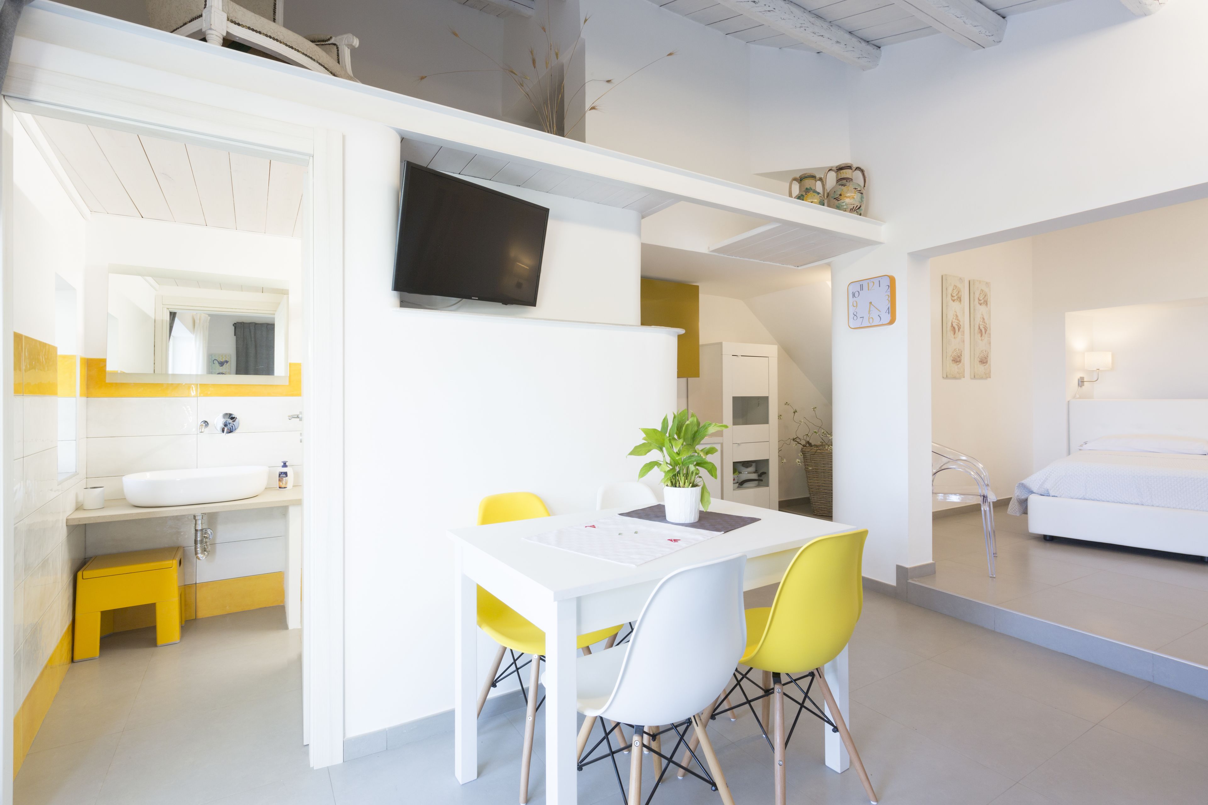 A modern, well-lit room with a white sink, yellow chairs, a white table, and a bed with white bedding.