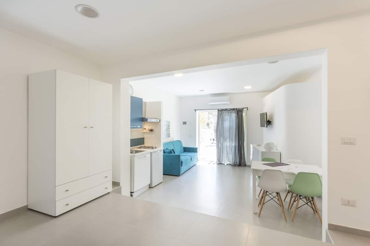 A modern kitchen with white cabinets, a dining area with a table and chairs, and a living area with a sofa and curtains.