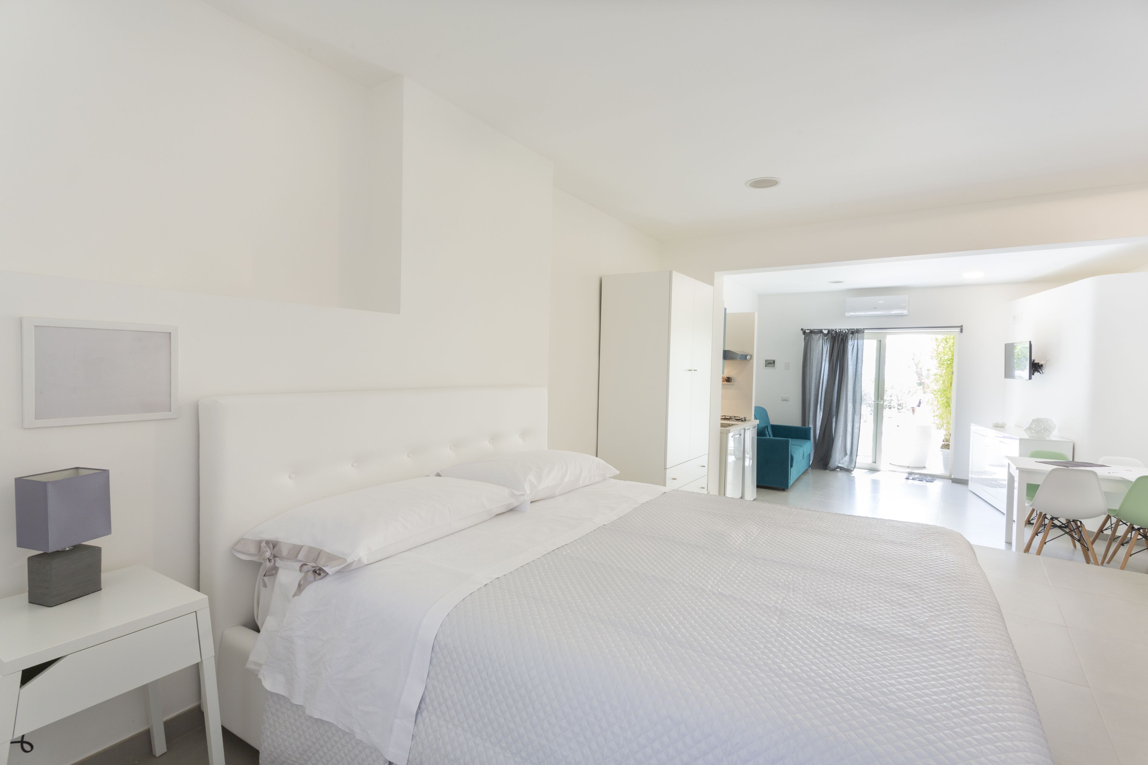 A modern bedroom with a large bed, a bedside table with a lamp, and a view of the bathroom and balcony.