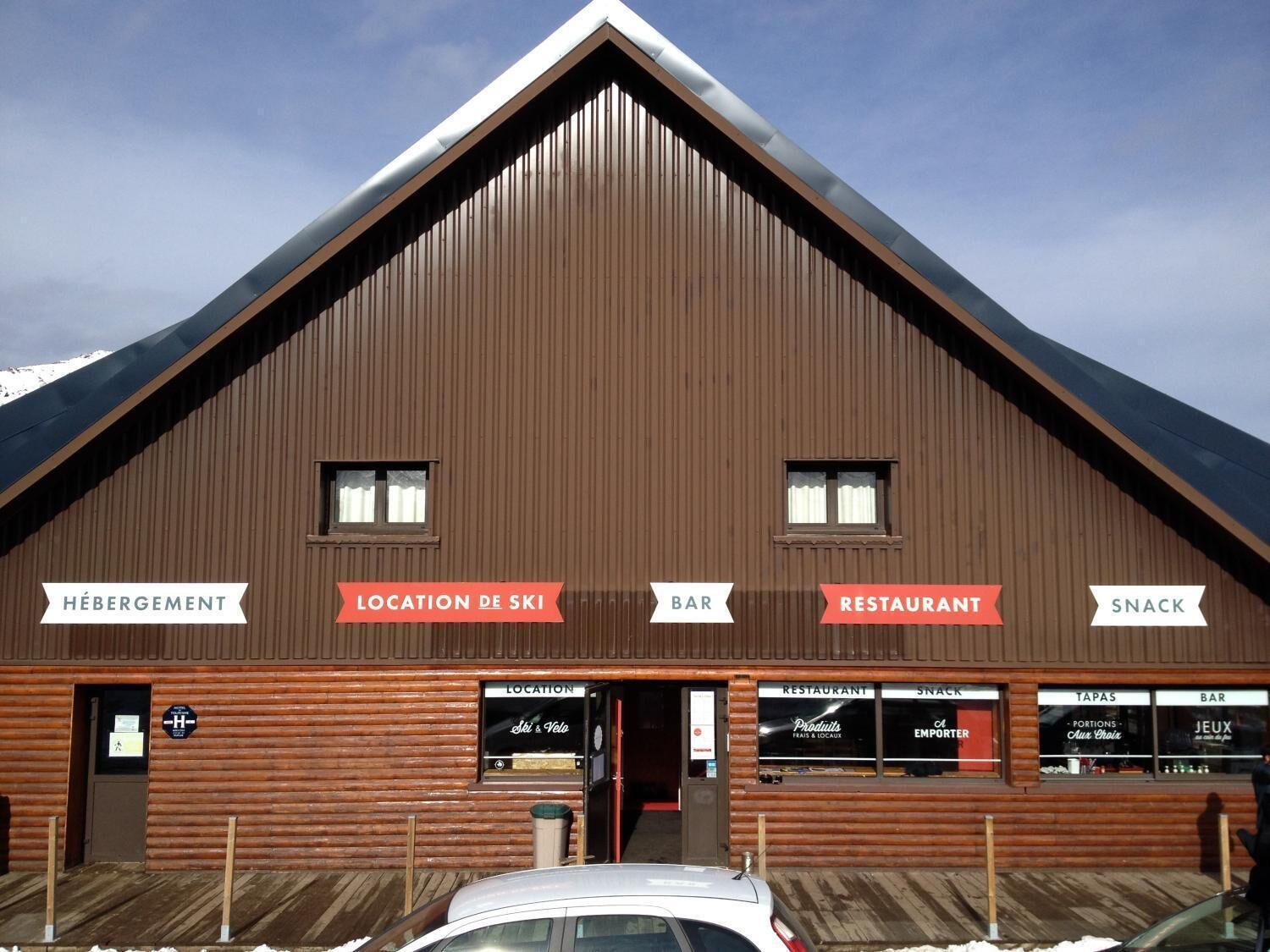 Edificio de madera con letreros que indican 'Hébergement', 'Location de Ski', 'Bar', 'Restaurant' y 'Snack'.