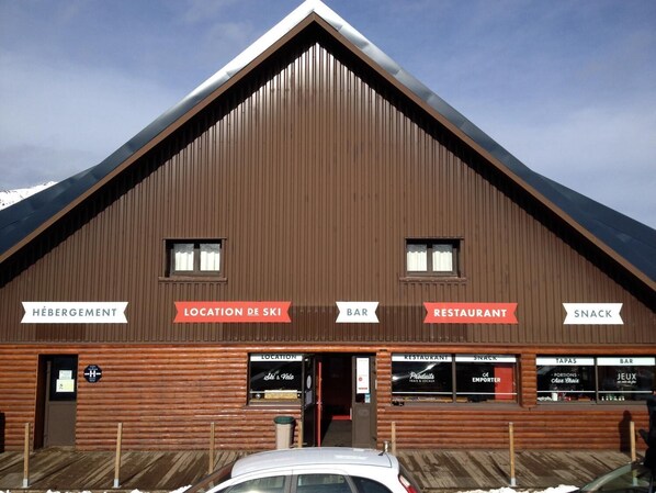 Edificio de madera con letreros que indican 'Hébergement', 'Location de Ski', 'Bar', 'Restaurant' y 'Snack'.