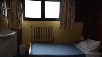Una stanza piccola e scarsamente illuminata con un letto singolo, un comodino, una finestra con tende, un termosifone e un lavandino.