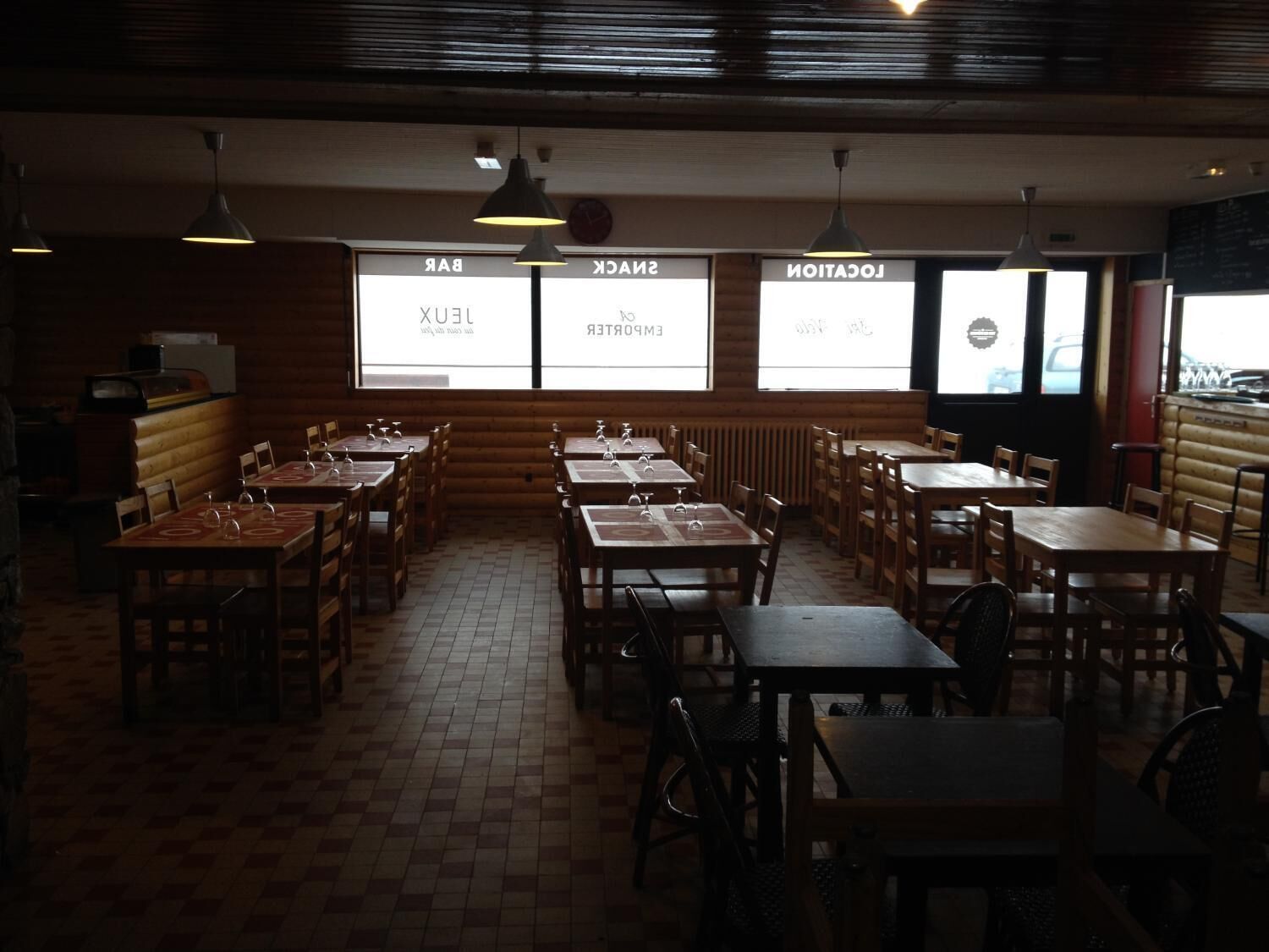 Un restaurant avec des tables et des chaises en bois, un sol carrelé et de grandes fenêtres sur lesquelles sont affichés les menus.