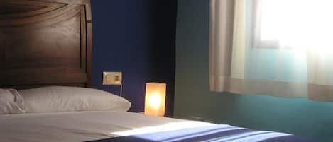 Una cama bien hecha con sábanas blancas, cabecera de madera, lámpara de noche y una ventana con cortinas translúcidas que dejan entrar la luz natural.