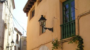 A narrow street with a building featuring a green window, a stone archway, and a sign with a blue 'H' indicating a hotel. - Cal Maginet Hotel (Vilaverd)