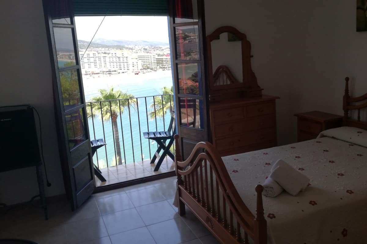 A bedroom with a bed, a wooden dresser, a nightstand, and a balcony with a view of a beach and buildings.
