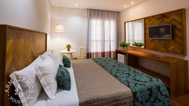 A hotel room with a bed, a desk, a TV, and a window with curtains. - Pousada Armazem 26 (Tiradentes)