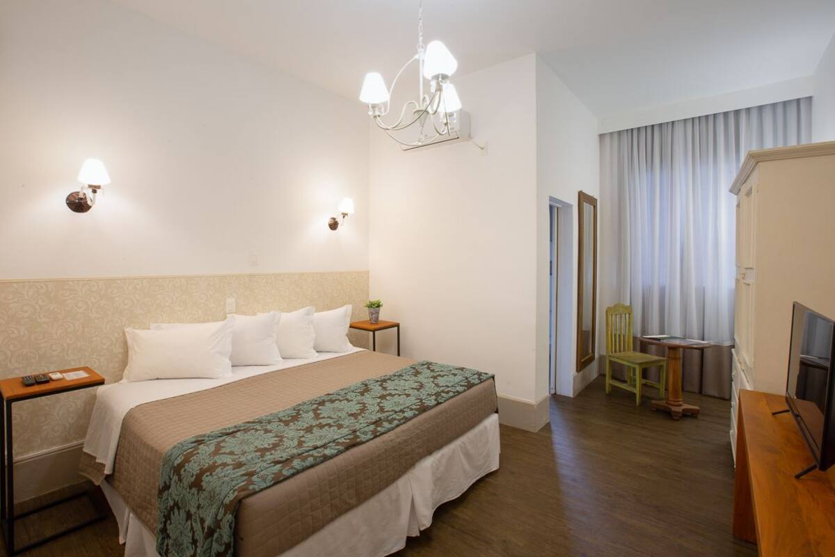 A hotel room with a large bed, a bedside table with a lamp, a small table with a chair, a TV, and a window with curtains.