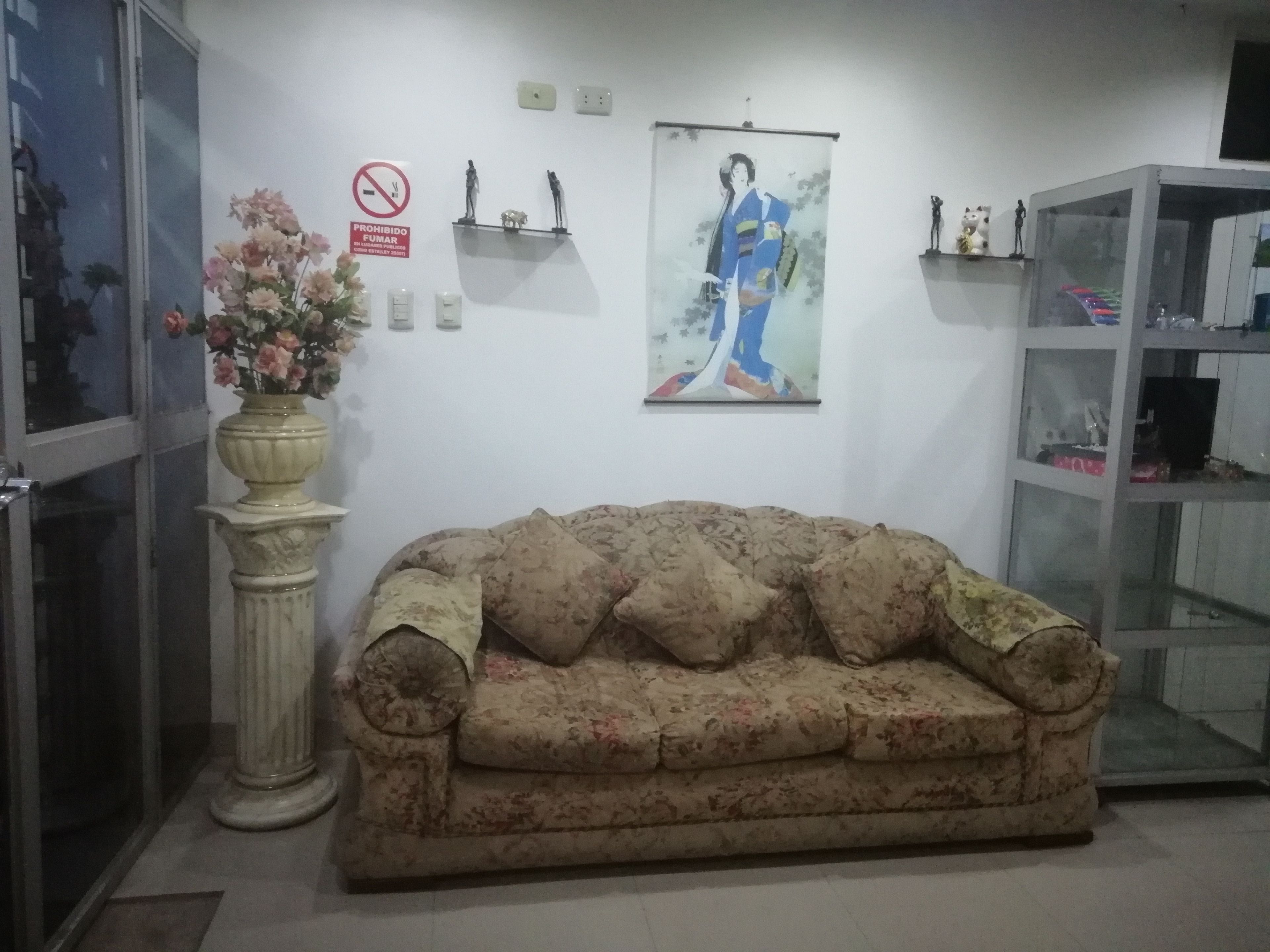 A room with a floral-patterned sofa, a vase with flowers, a framed picture of a person in traditional attire, and a cabinet with various items.