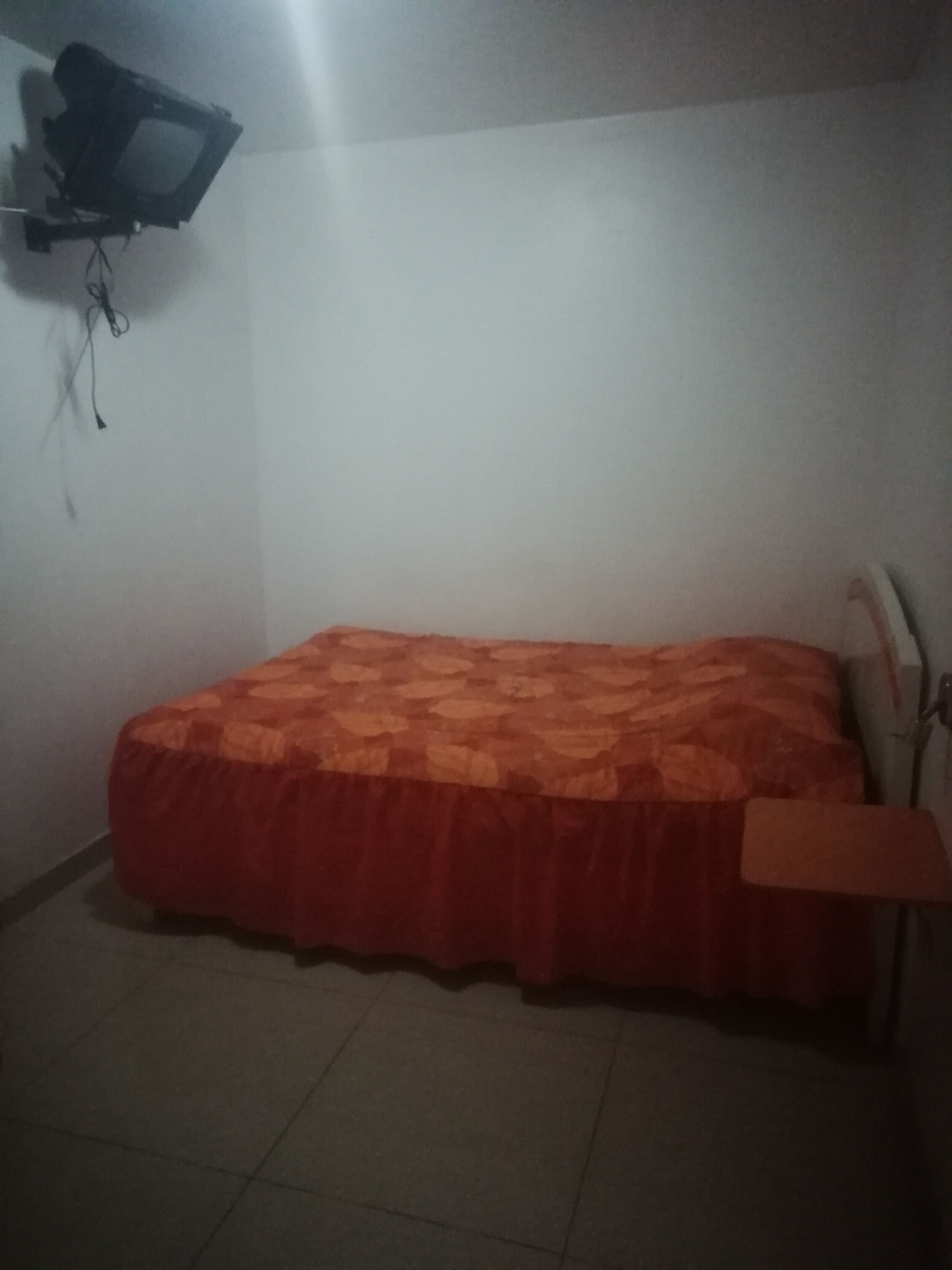 Una habitación pequeña y tenuemente iluminada con una cama individual, una silla y un televisor fijado a la pared.