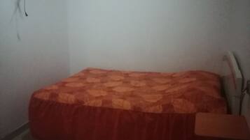 Una habitación pequeña y tenuemente iluminada con una cama individual, una silla y un televisor fijado a la pared.