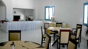 A dining area with round tables and chairs, a long table with a white tablecloth, and a window with blue frames. - Captains Rock (Paros)