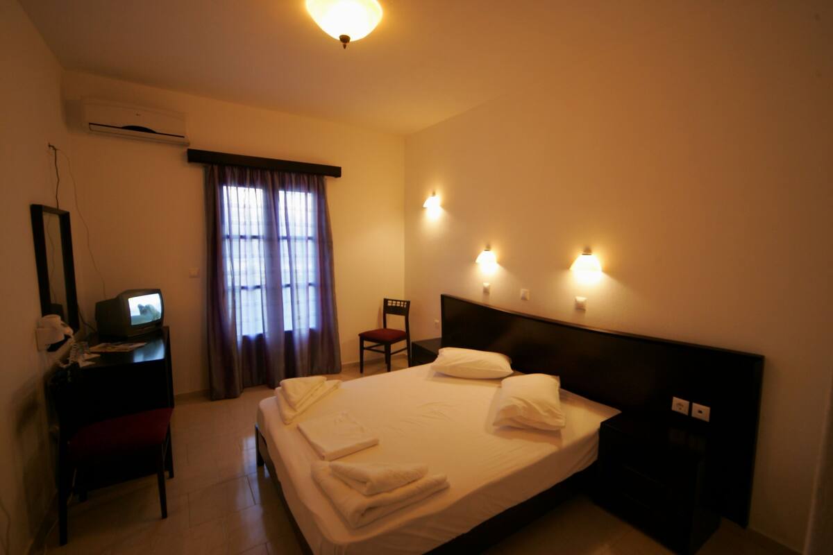 A hotel room with a bed, a desk with a television, a chair, and a window with curtains.