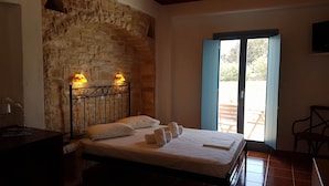 A bedroom with a stone wall, a bed with white linens, two bedside lamps, and a view through a glass door. - Captains Rock (Paros)