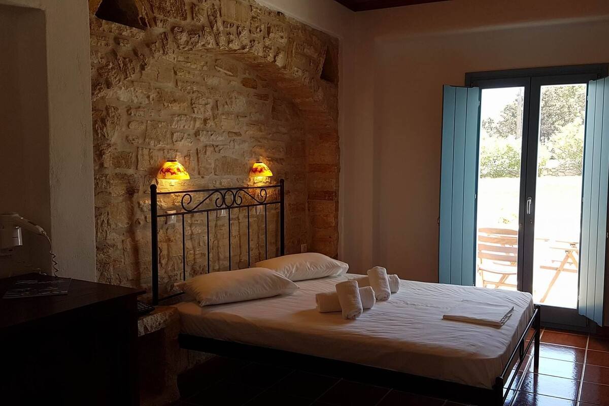 A bedroom with a stone wall, a bed with white linens, two bedside lamps, and a view through a glass door.