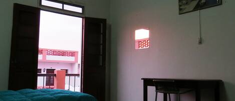 A room with two beds, a desk, a small table, a wall-mounted light, and a window with a view of buildings outside.