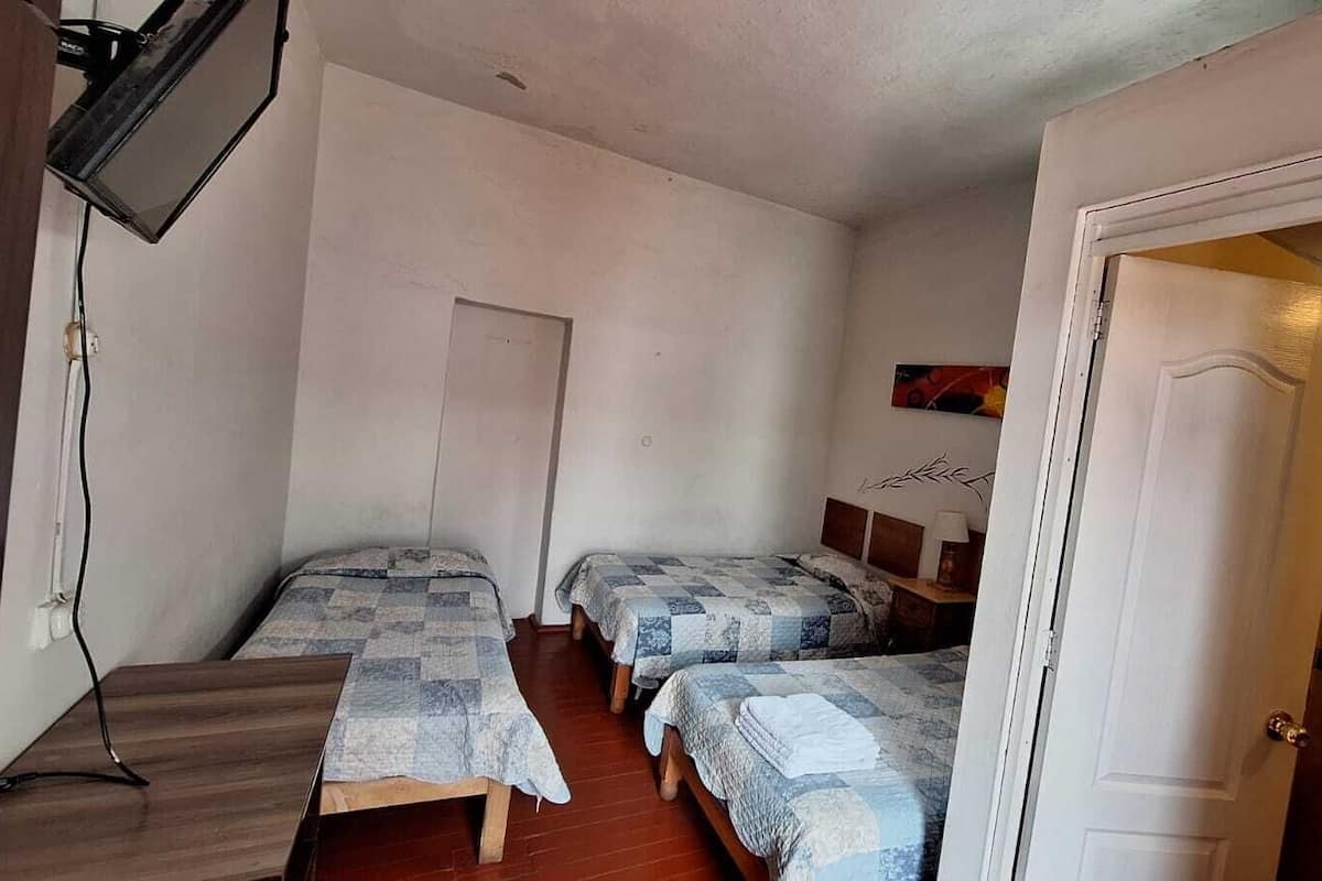 A small, compact room with three single beds, a wooden desk, a television mounted on the wall, and a door leading to a bathroom.