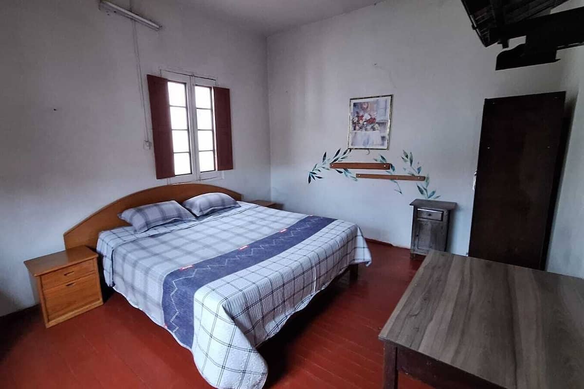 A bedroom with a single bed, wooden headboard, a nightstand, a small table, and a window with shutters.