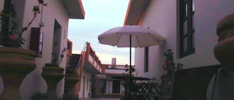A narrow outdoor corridor with potted plants, a table with chairs, and a large umbrella.