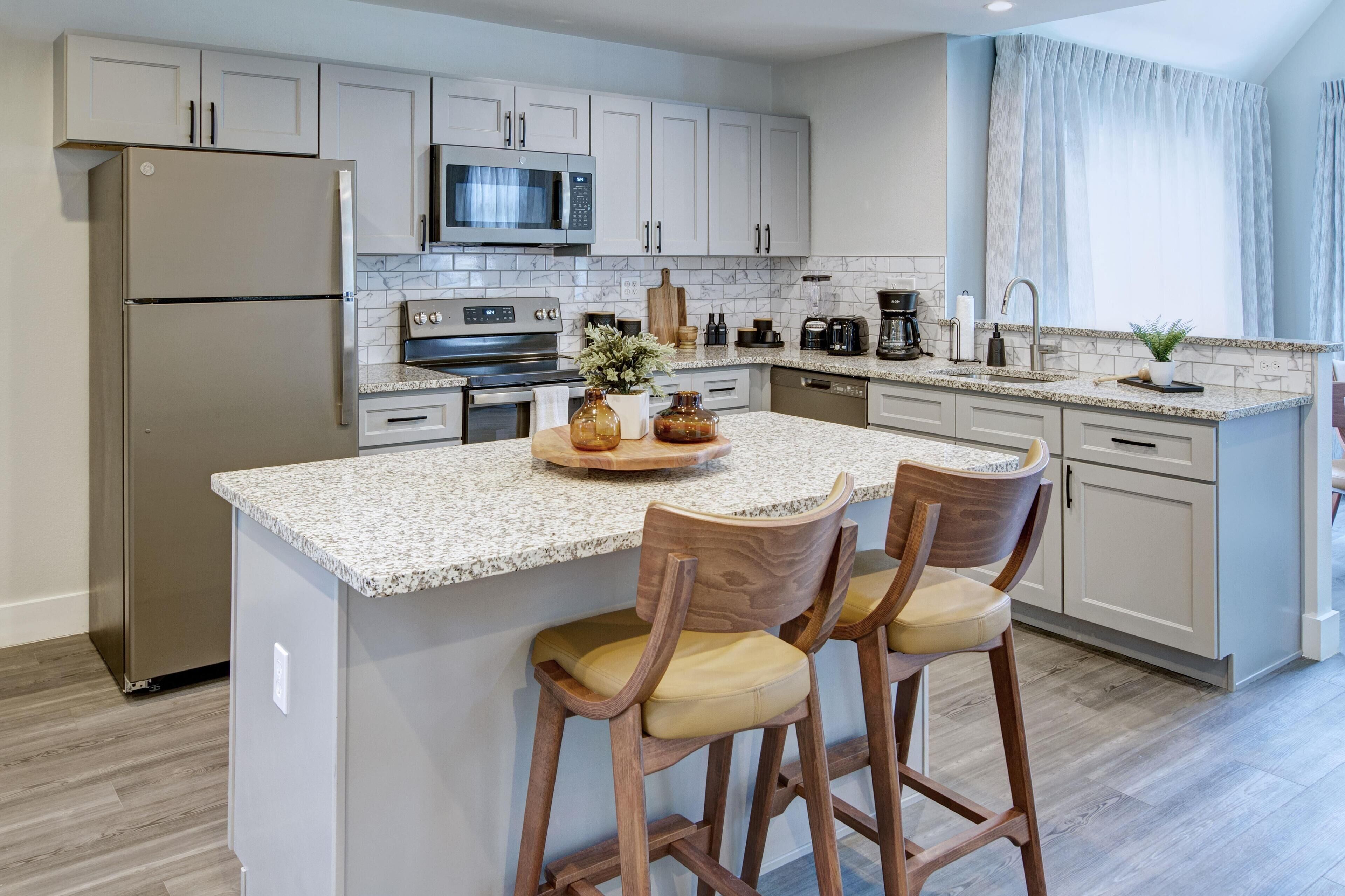 A modern kitchen with a central island, bar stools, a refrigerator, microwave, oven, and a coffee maker.