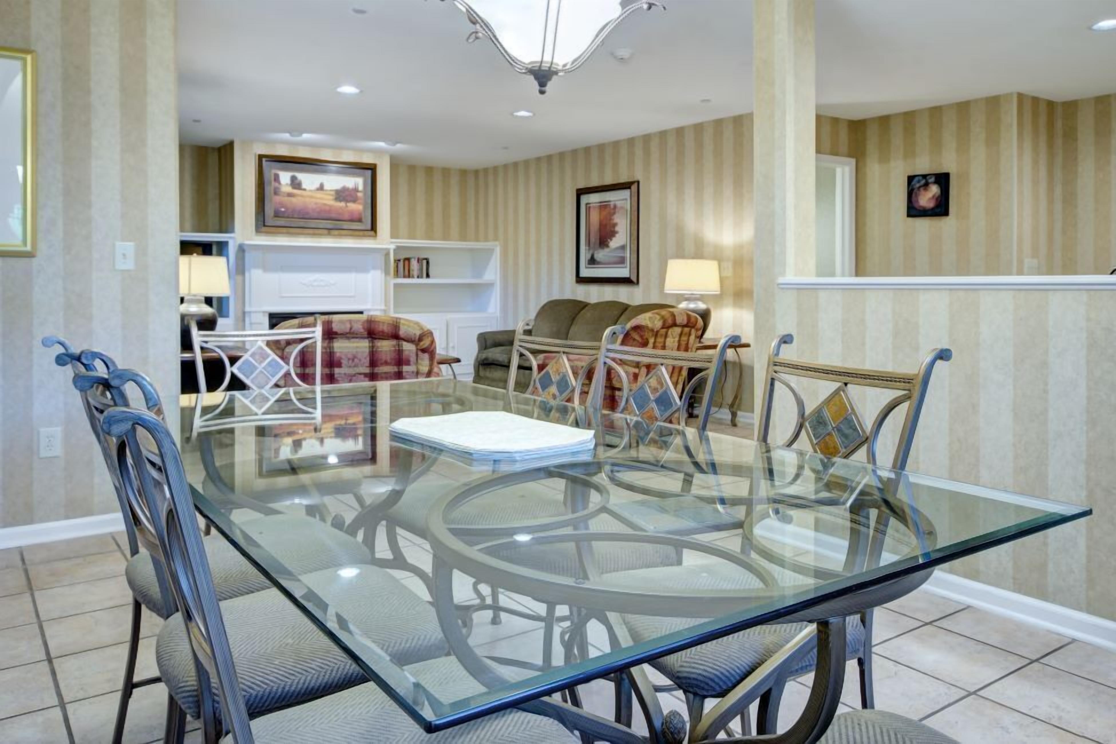 A dining area with a glass table and metal chairs, a sofa, and a fireplace in the background.
