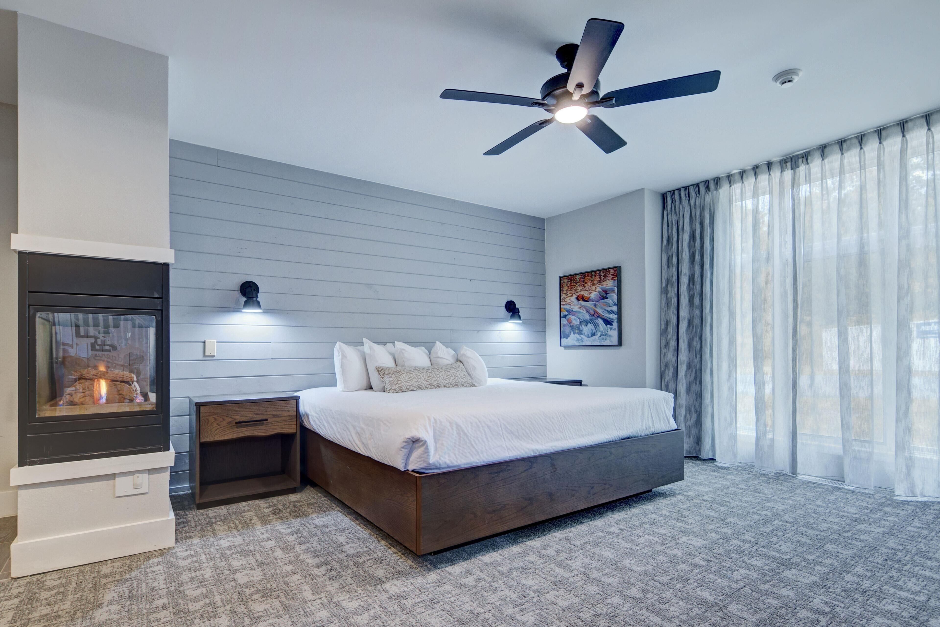 A modern bedroom with a large bed, a built-in fireplace, a ceiling fan, and a large window with sheer curtains.