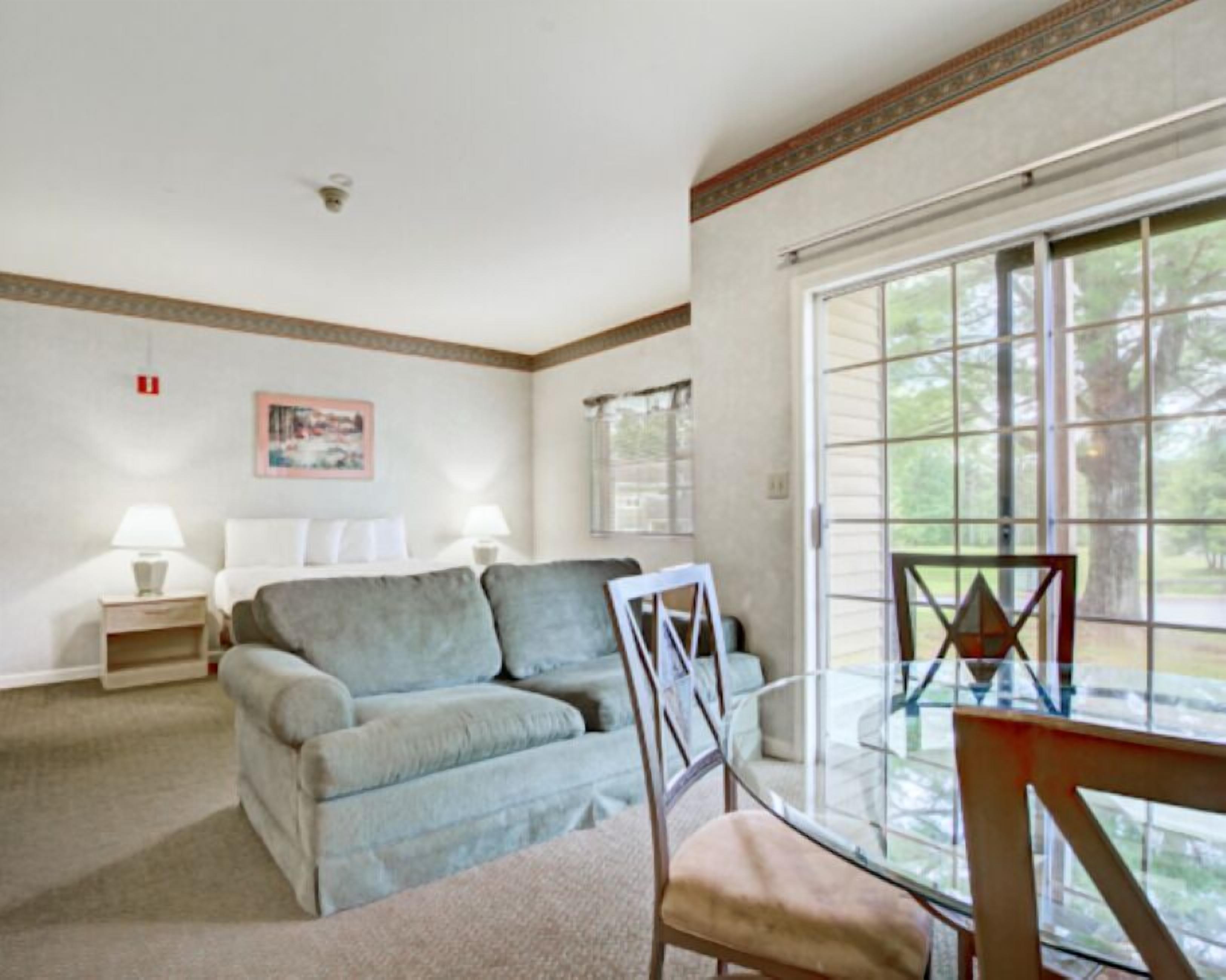 A room with a bed, a sofa, a glass table, and a chair. There is a window with a view of trees.