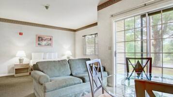 A room with a bed, a sofa, a glass table, and a chair. There is a window with a view of trees.