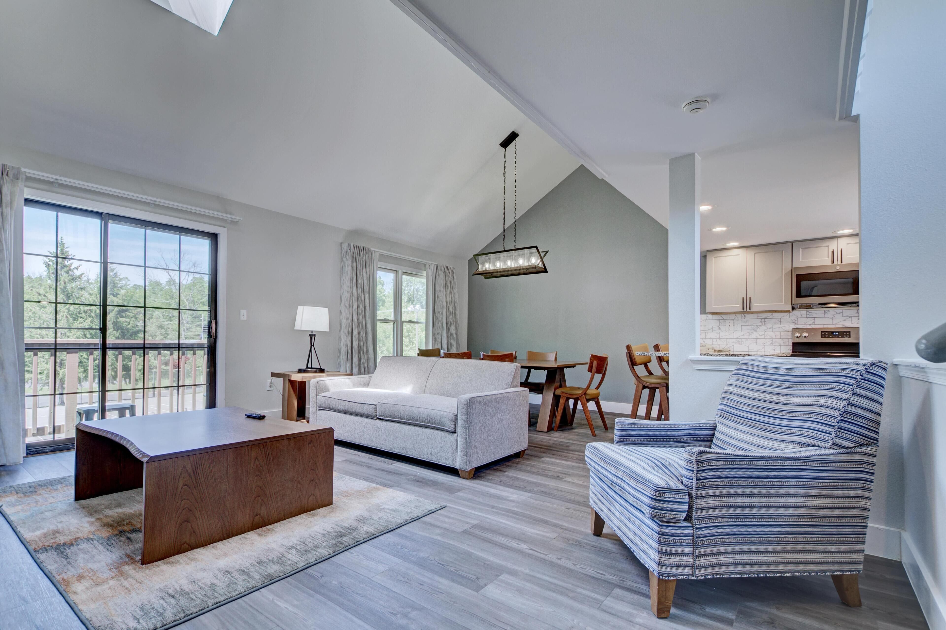 A modern living room with a sofa, a coffee table, a striped armchair, and a dining area with a table and chairs.