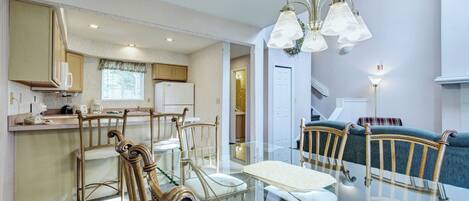 A dining area with a glass table and chairs, a kitchen in the background, and a staircase leading to another level.