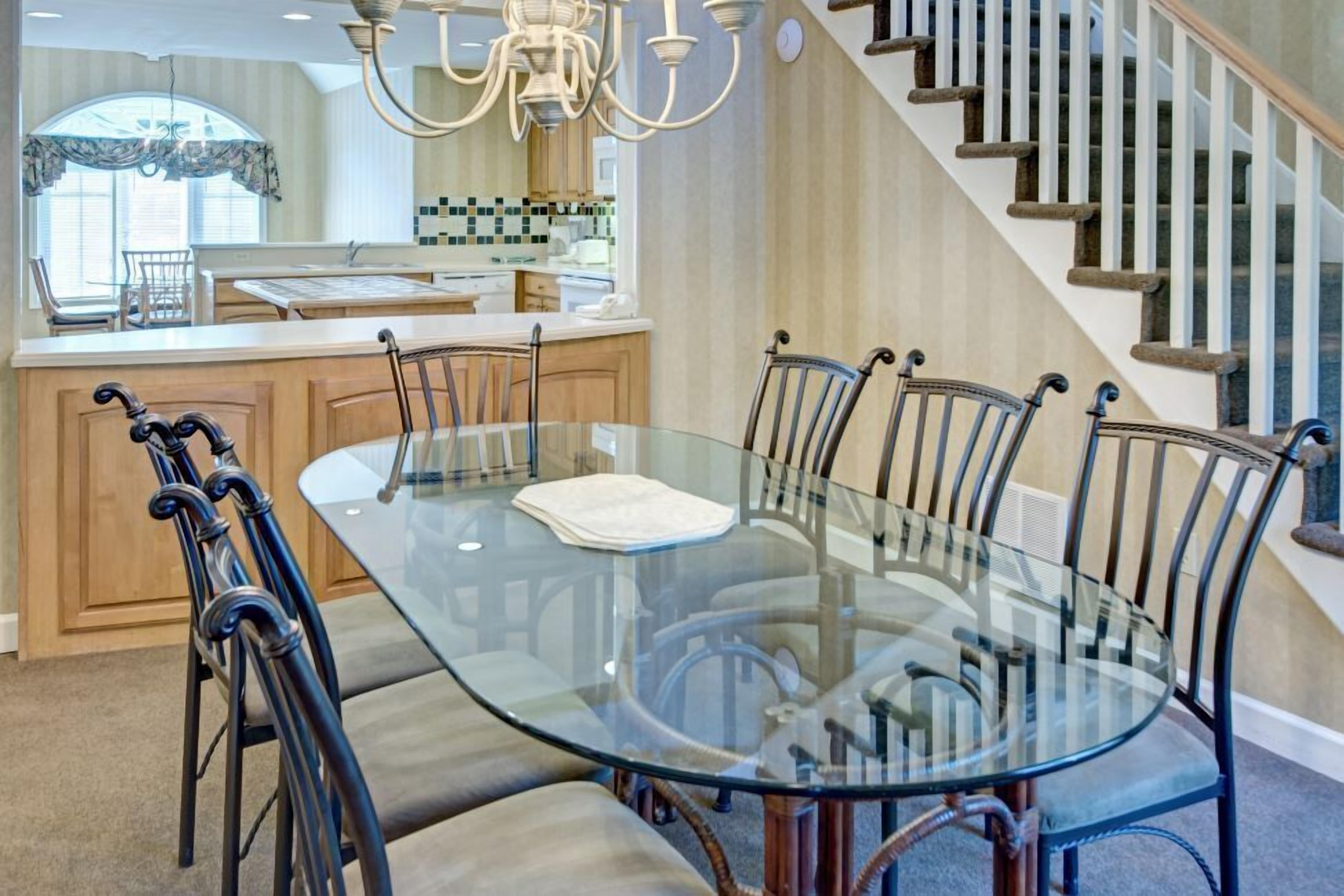 Un coin repas avec une table et des chaises en verre, un lustre, une cuisine en arrière-plan et un escalier.