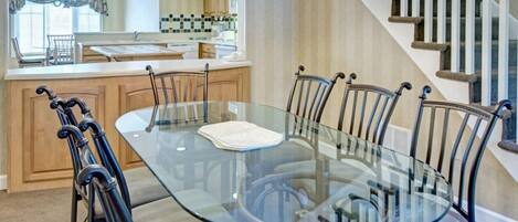 Un coin repas avec une table et des chaises en verre, un lustre, une cuisine en arrière-plan et un escalier.