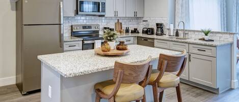 A modern kitchen with a central island, bar stools, a refrigerator, microwave, oven, and a coffee maker.