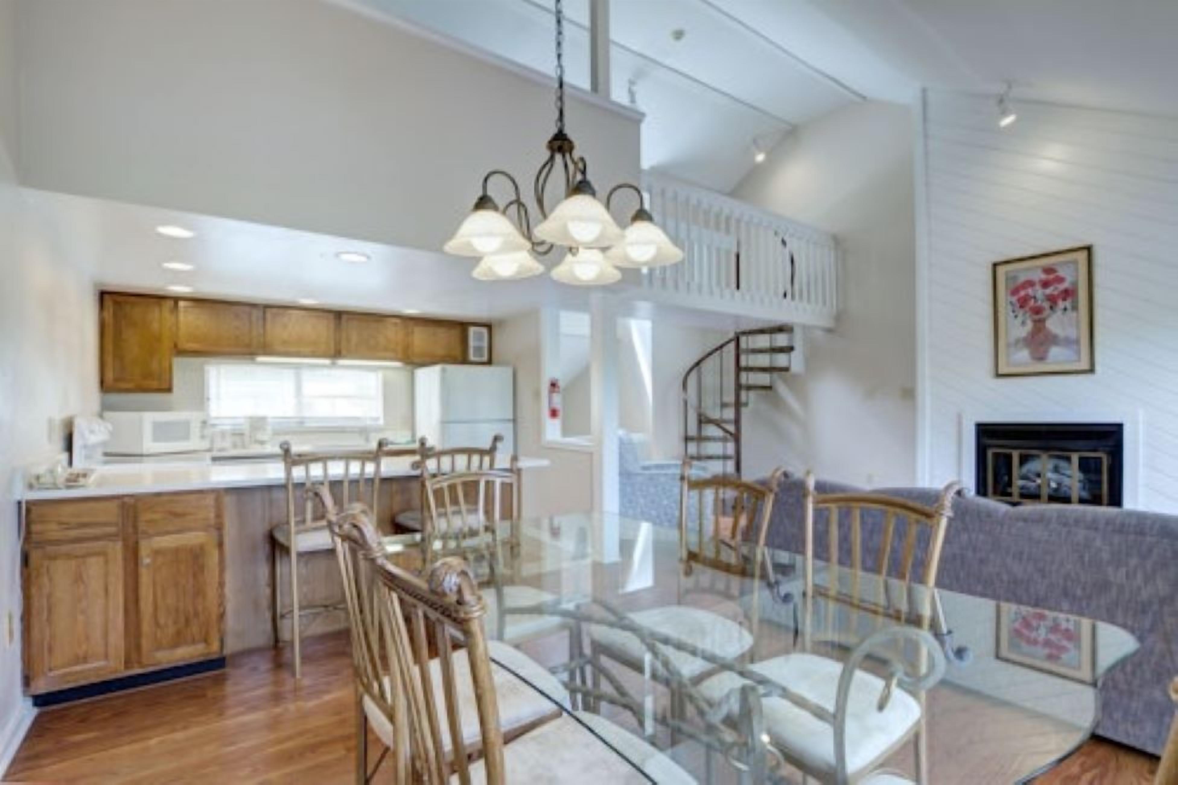 Cocina amplia con armarios de madera, comedor con mesa y sillas de vidrio, y una escalera que lleva a un nivel superior.