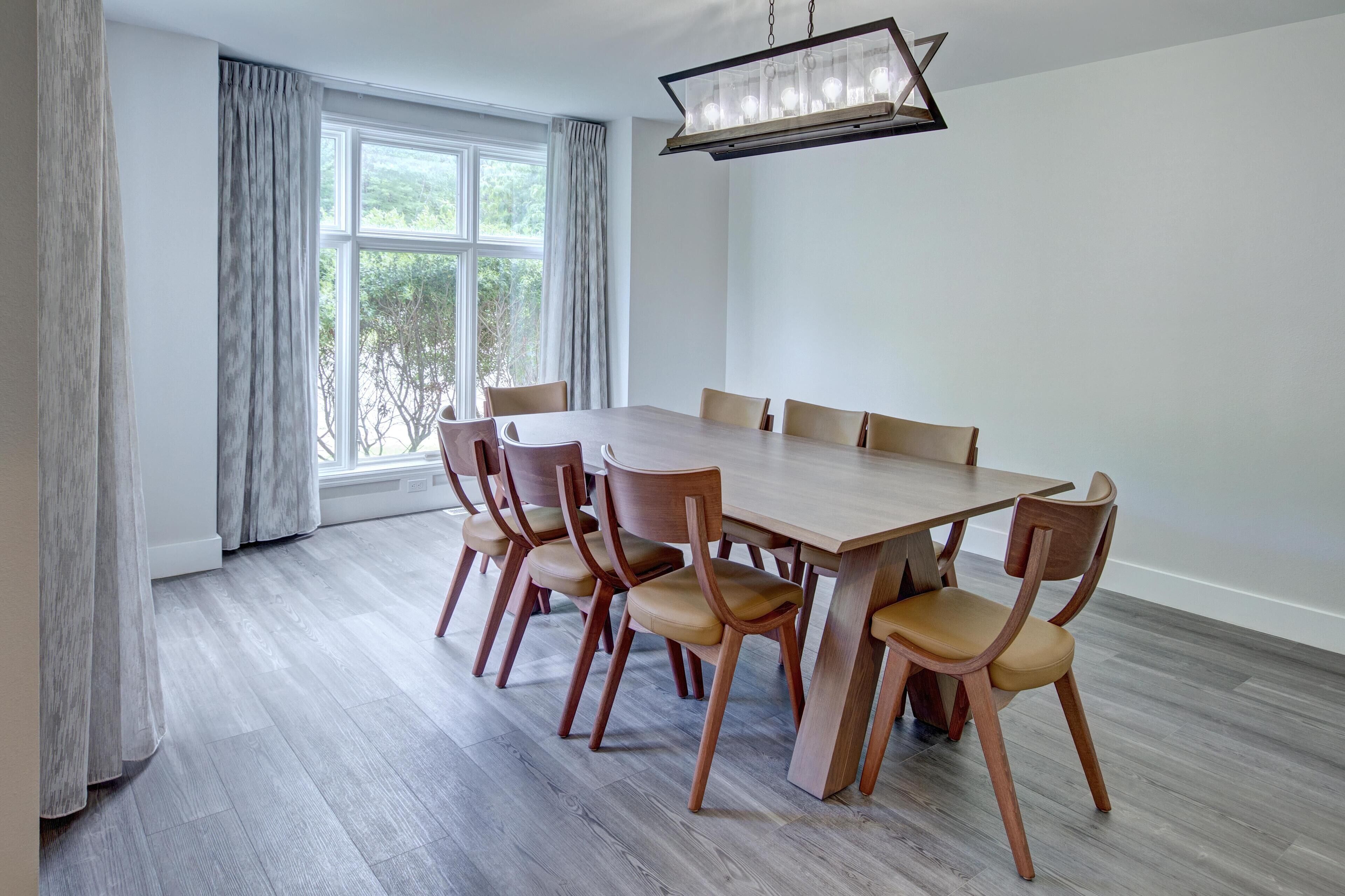 Un comedor con mesa y sillas de madera, ventanas amplias con cortinas y una lámpara colgante.