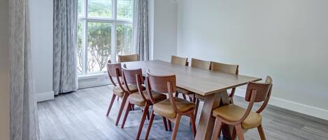 Un comedor con mesa y sillas de madera, ventanas amplias con cortinas y una lámpara colgante.