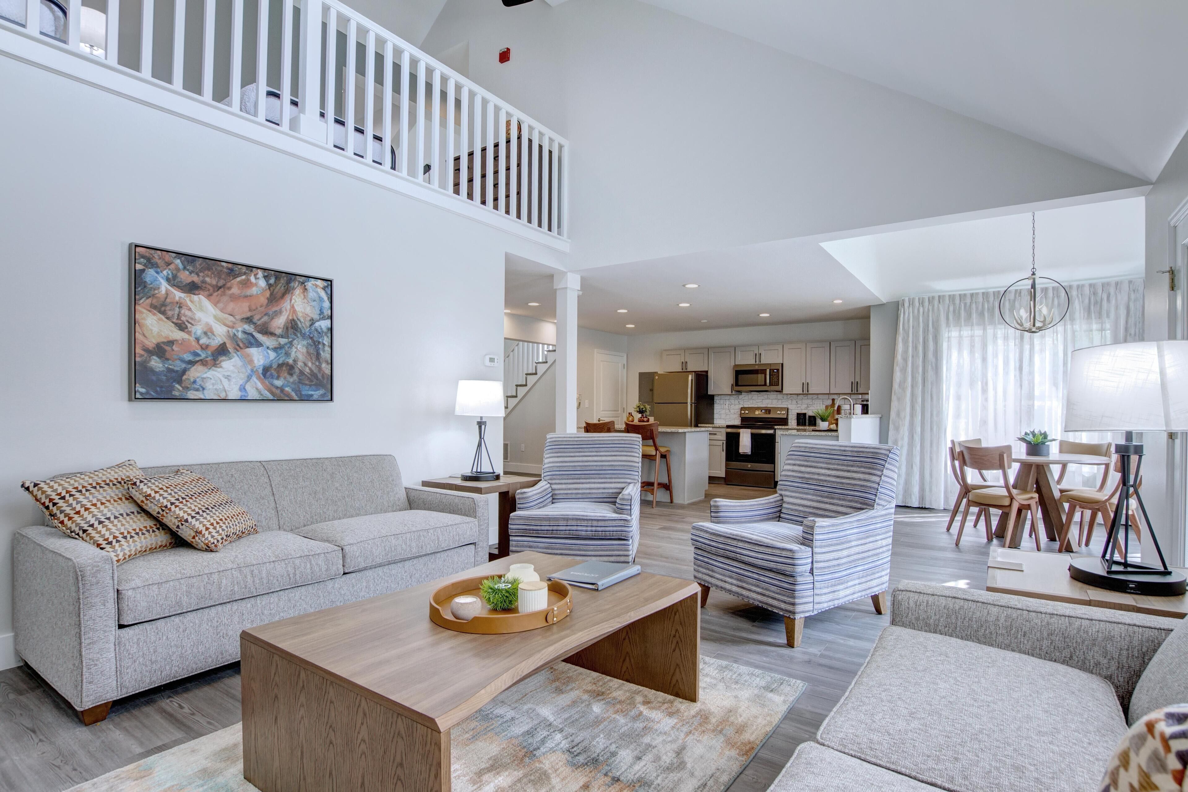 A spacious living area with a high ceiling, a staircase, and a kitchen in the background.