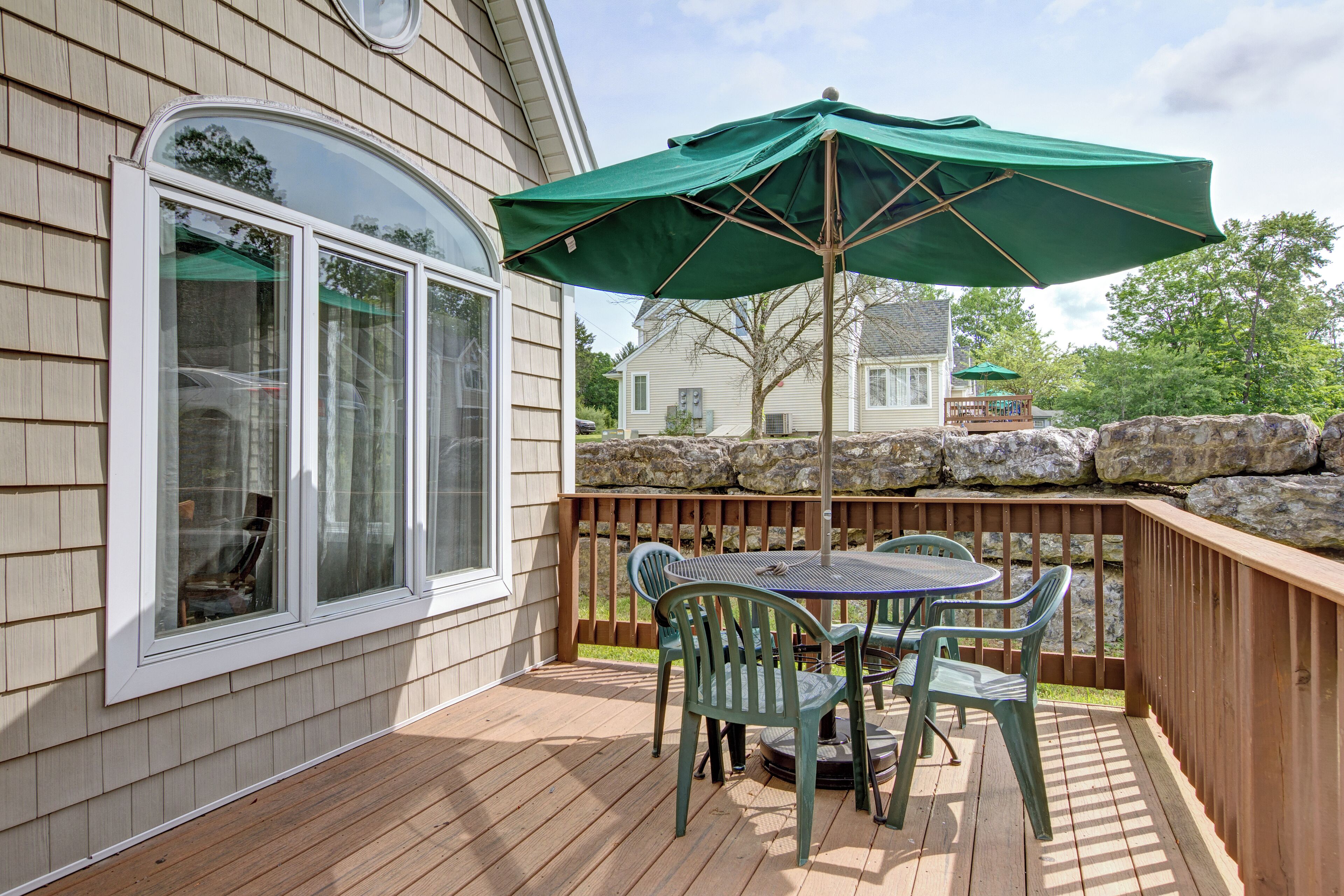 A wooden deck with a table and chairs, a green umbrella, and a stone wall.