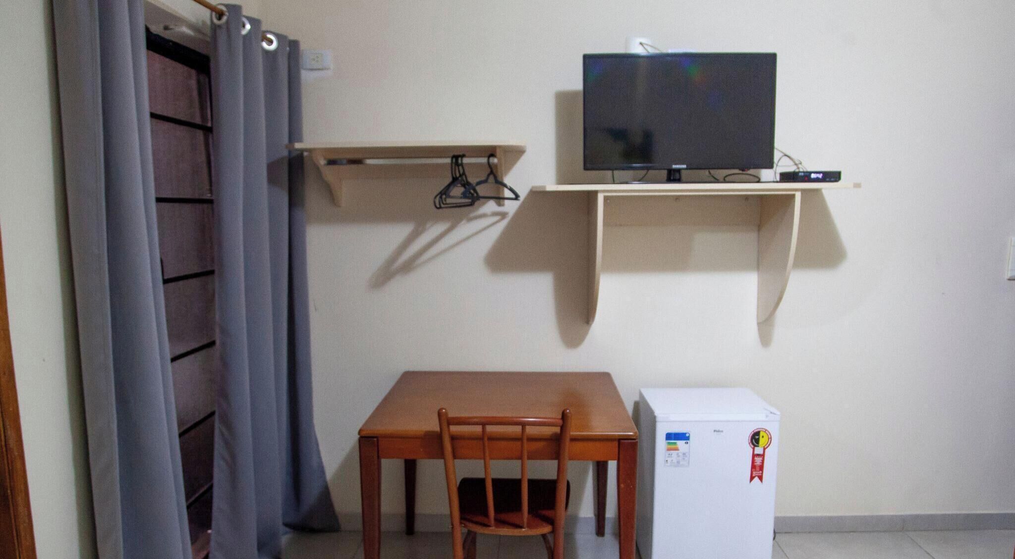 Een kleine kamer met een houten tafel, een stoel, een televisie op een plank en een koelkast.