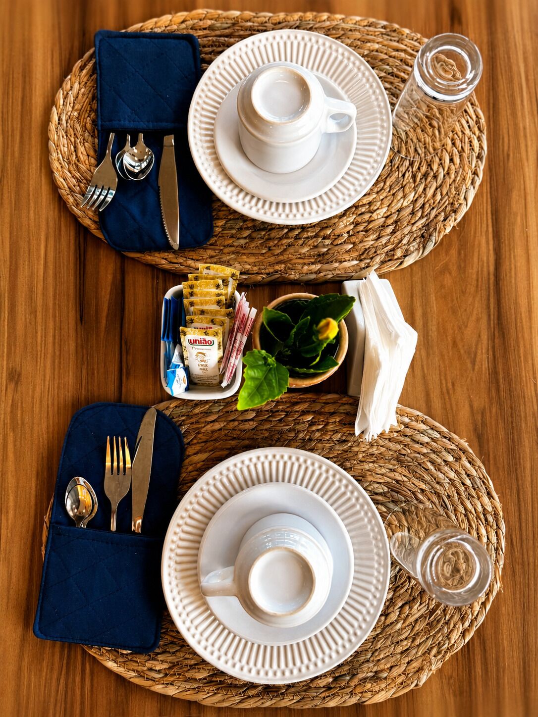 A neatly arranged breakfast setting with two woven placemats, white plates, cups, utensils, and condiments on a wooden table.