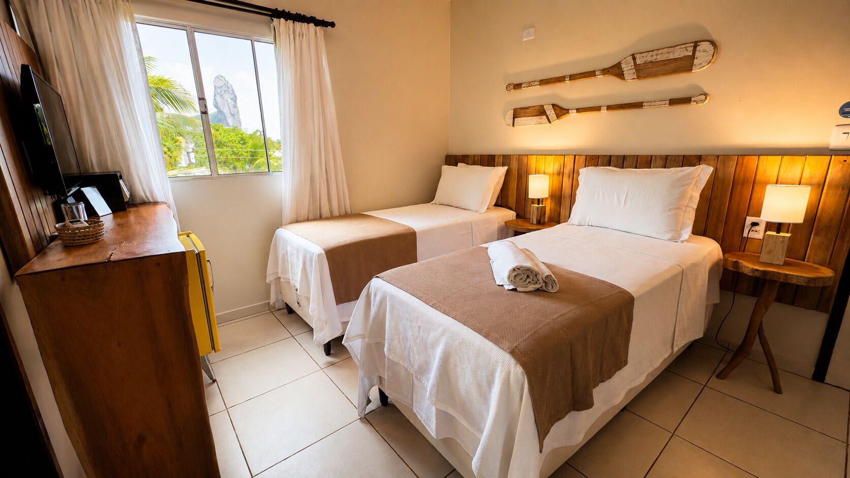 A hotel room with two beds, a wooden desk, a television, and a view of greenery outside.
