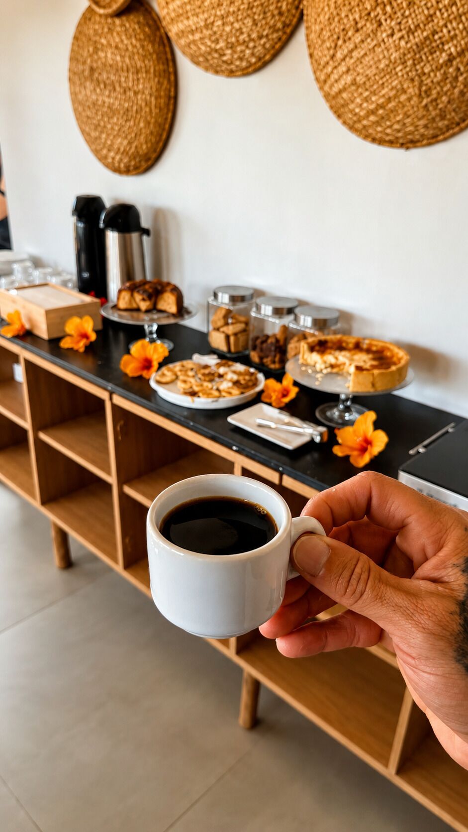 Una mano sosteniendo una taza de café, una mesa de buffet con diferentes platillos y una cafetera al fondo.