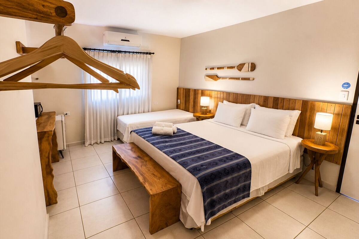 Ein Hotelzimmer mit einem großen Bett, einer hölzernen Bank, einem Holzaufhänger, einem Nachttisch mit Lampe und einer Wand-Klimaanlage.