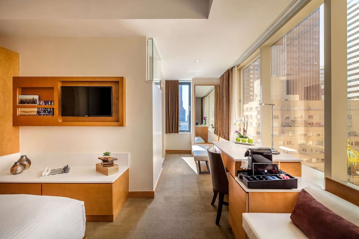 Ein modernes Hotelzimmer mit einem großen Bett, einem Schreibtisch mit Kaffeemaschine, einer Sitzecke und Blick auf die Stadt durch große Fenster.