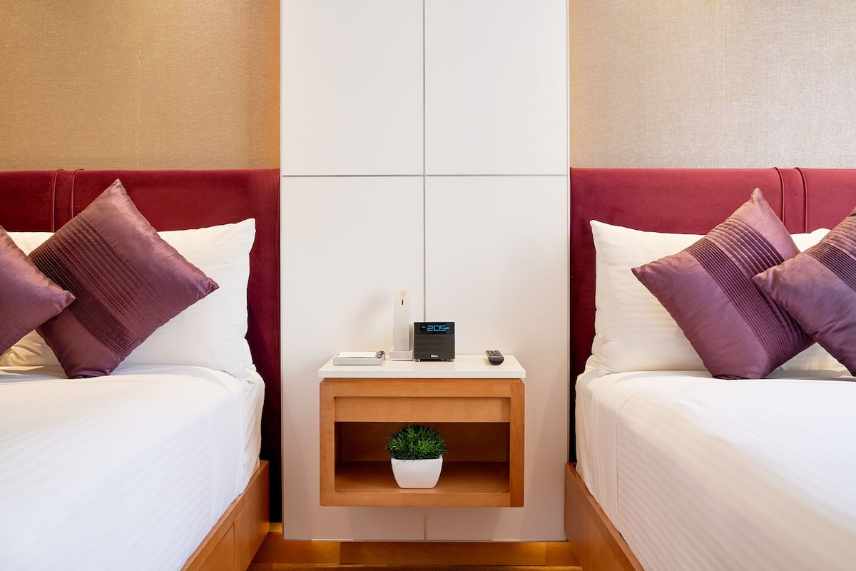 Ein Hotelzimmer mit zwei Betten, beide mit rotem Kopfteil, weißer Bettwäsche und violetten Kissen. Ein kleiner Nachttisch aus Holz mit einer Uhr, einer Fernbedienung und einer Topfpflanze.