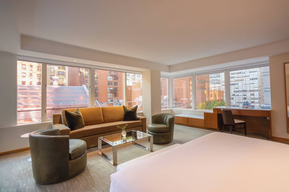 Ein modernes Hotelzimmer mit einem großen Bett, einer Sitzecke, einem Schreibtisch und Blick auf die Stadt durch große Fenster.