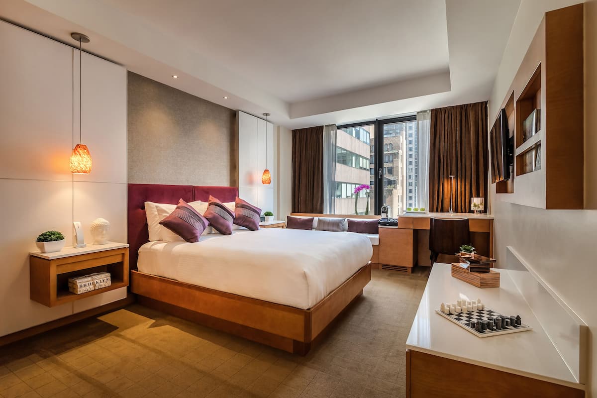 Ein modernes Hotelzimmer mit einem großen Bett, Nachttischen, einem Schreibtisch mit Schachbrett und Blick auf die Stadt durch große Fenster.