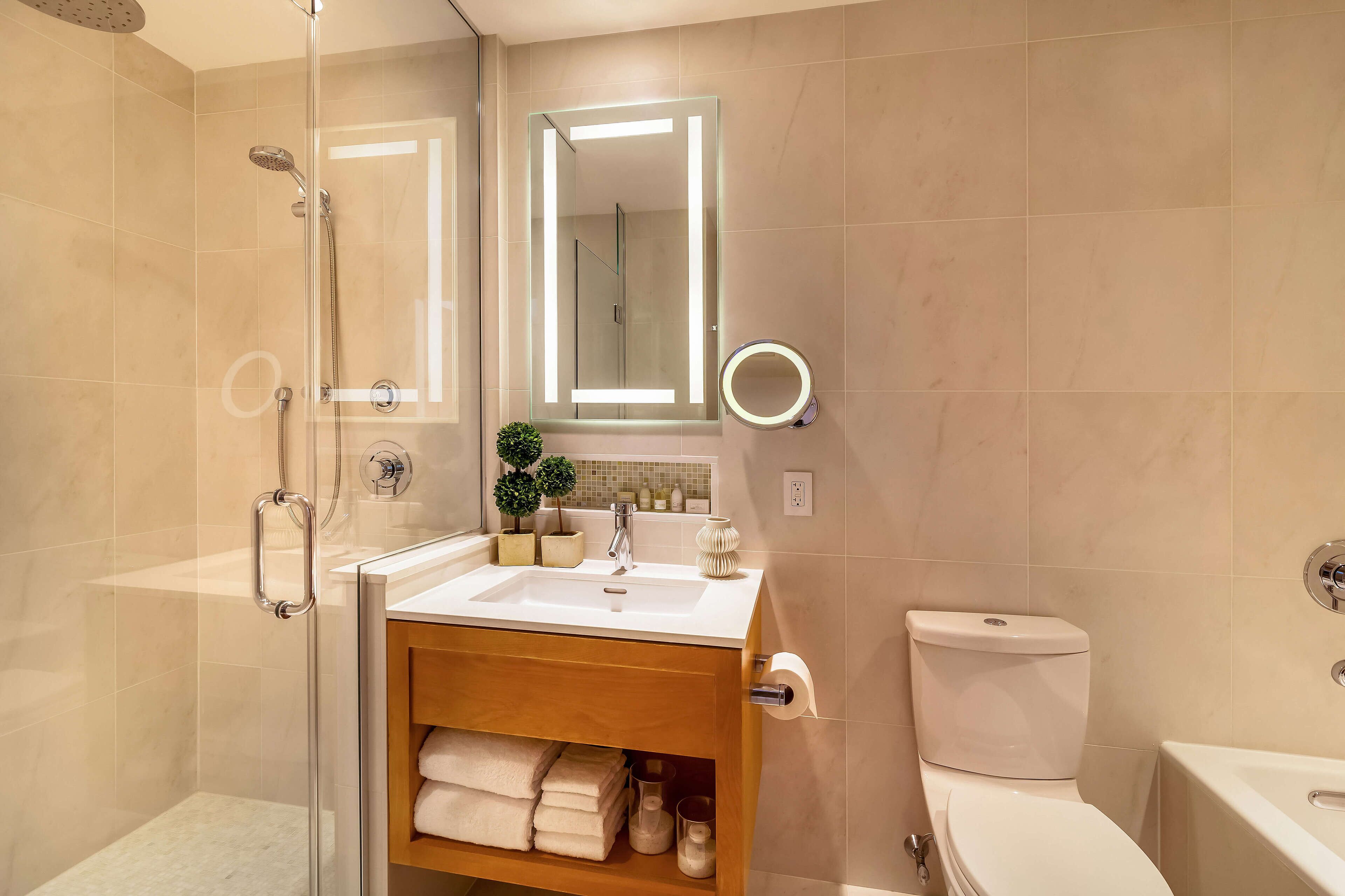 Un baño moderno con cabina de ducha de vidrio, tocador con lavabo, inodoro y espejo con iluminación integrada.