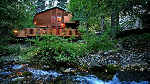Una baita di legno con una veranda, immersa in una foresta e accanto a un ruscello.