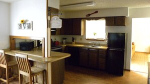 A compact kitchen with wooden cabinets, a built-in microwave, and a refrigerator. There is a small island with bar stools and a window providing natural light.